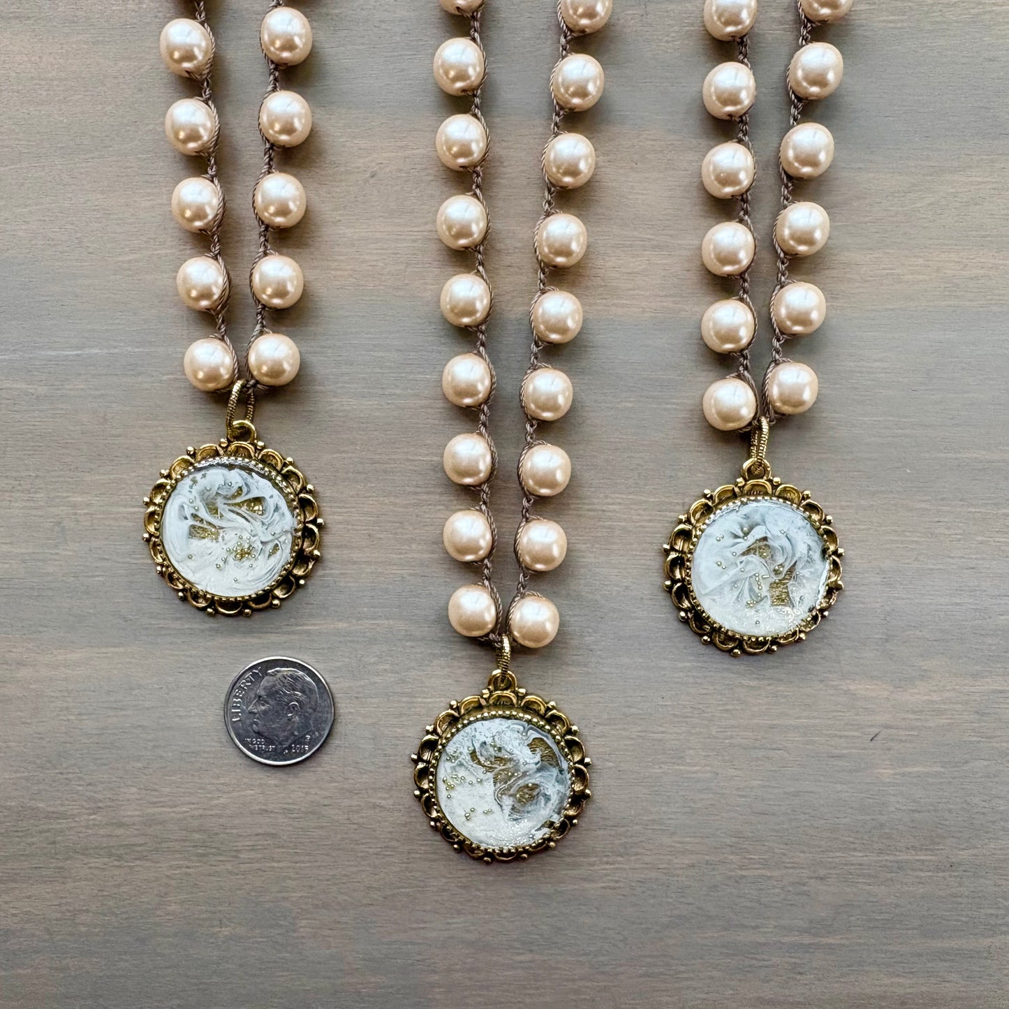 Two pearl necklaces with gold pendants on a wooden surface, one with a coin for scale.