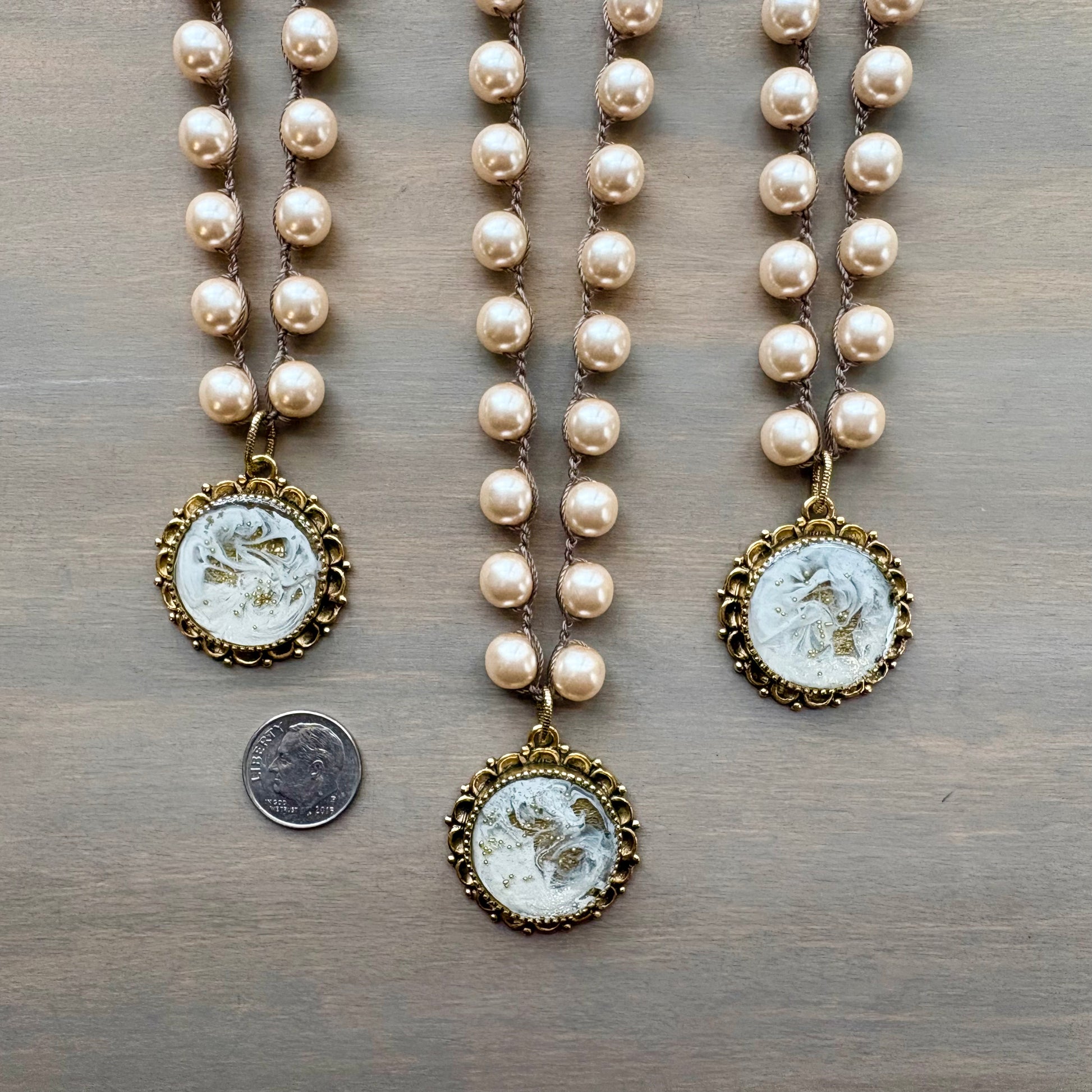 Two pearl necklaces with gold pendants on a wooden surface, one with a coin for scale.