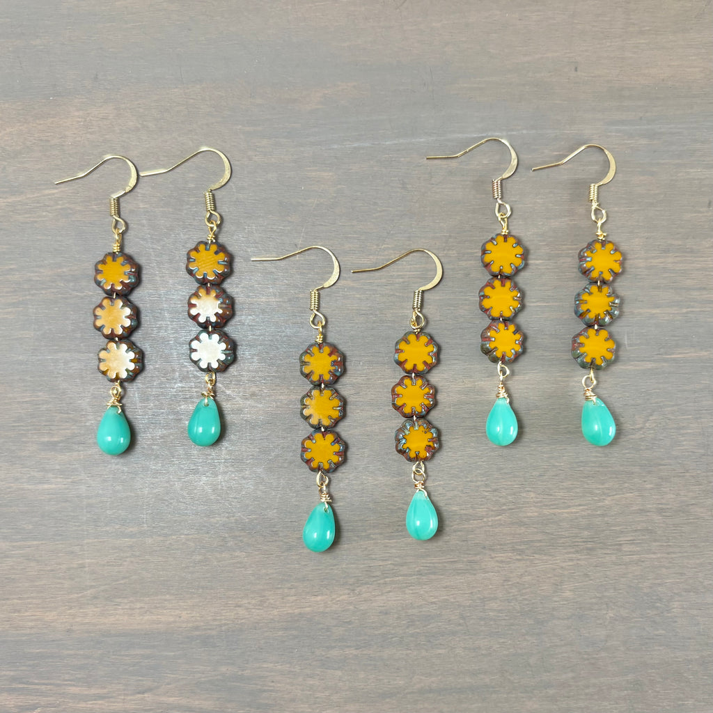 Three pairs of dangling earrings laid flat on a gray wood surface. Each pair has gold-tone fishhook ear wires, four stacked flower-shaped beads in yellow, brown, and white, and a turquoise teardrop bead at the bottom.