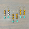 Three pairs of dangling earrings laid flat on a gray wood surface. Each pair has gold-tone fishhook ear wires, four stacked flower-shaped beads in yellow, brown, and white, and a turquoise teardrop bead at the bottom.