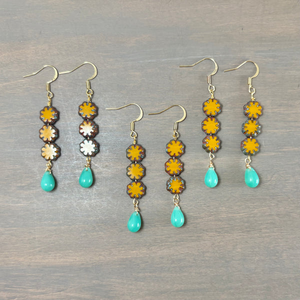 Three pairs of dangling earrings laid flat on a gray wood surface. Each pair has gold-tone fishhook ear wires, four stacked flower-shaped beads in yellow, brown, and white, and a turquoise teardrop bead at the bottom.