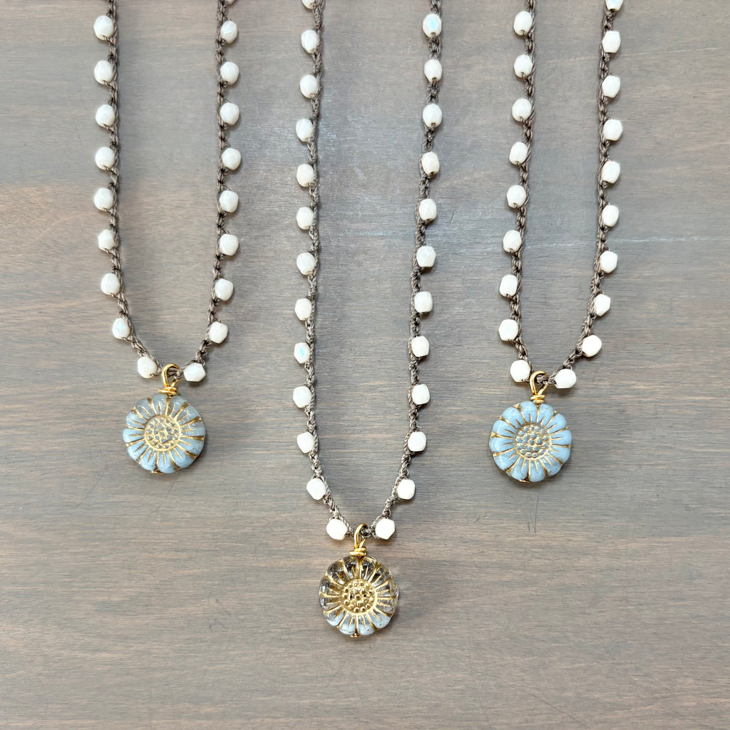 Three beaded necklaces with round flower pendants are arranged on a gray wood background. The necklaces feature white faceted beads on dark metal chains and have light blue and gold flower charms.