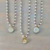 Three beaded necklaces with round flower pendants are arranged on a gray wood background. The necklaces feature white faceted beads on dark metal chains and have light blue and gold flower charms.