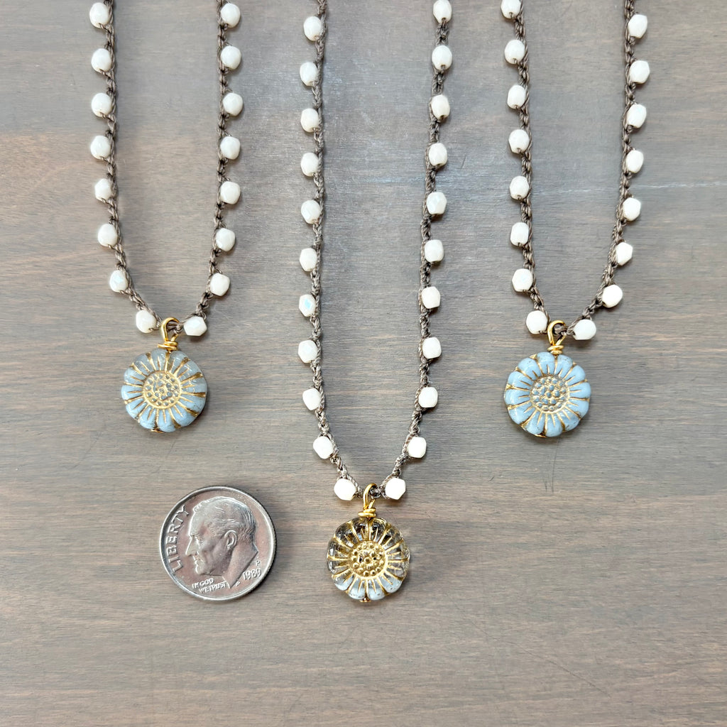 Three beaded necklaces with round flower-shaped pendants are arranged on a gray wood surface, with a dime below the center pendant for scale. The necklaces have white faceted beads on dark chains and light blue and gold flower charms.