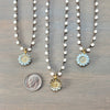 Three beaded necklaces with round flower-shaped pendants are arranged on a gray wood surface, with a dime below the center pendant for scale. The necklaces have white faceted beads on dark chains and light blue and gold flower charms.