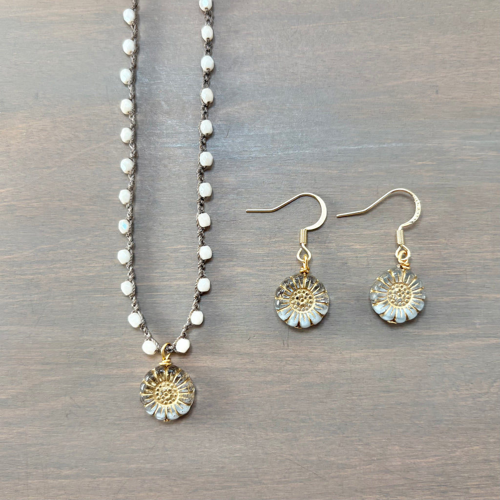 A jewelry set featuring a beaded necklace and matching dangle earrings laid flat on a dark wood background. The necklace and earrings have gold-tone metal with white bead details and light blue iridescent accents in the flower-shaped pendants.