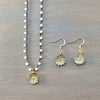A jewelry set featuring a beaded necklace and matching dangle earrings laid flat on a dark wood background. The necklace and earrings have gold-tone metal with white bead details and light blue iridescent accents in the flower-shaped pendants.