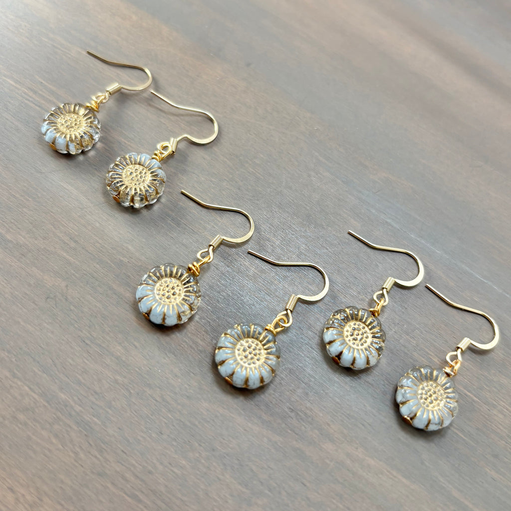 Four gold-tone dangle earrings with round white and gold flower-shaped charms, arranged diagonally on a dark wood surface.