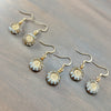 Four gold-tone dangle earrings with round white and gold flower-shaped charms, arranged diagonally on a dark wood surface.