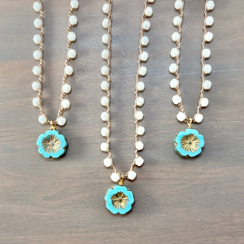 Three bohemian-style necklaces with gold chains, white seed beads, and turquoise flower pendants are arranged on a dark wood background.