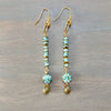 A pair of gold-tone dangle earrings with turquoise-blue beads, small gold spacer beads, and oval turquoise floral clusters ending in gold-tone teardrop charms, displayed on a gray wood background.