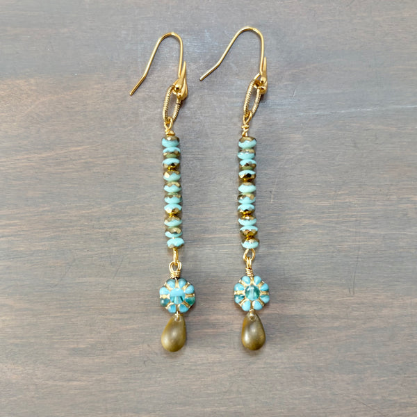 A pair of gold-tone dangle earrings with turquoise-blue beads, small gold spacer beads, and oval turquoise floral clusters ending in gold-tone teardrop charms, displayed on a gray wood background.