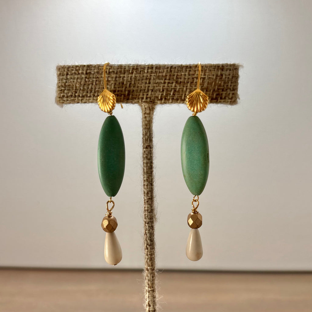 A pair of bohemian-style drop earrings displayed on a burlap-covered jewelry stand. The earrings feature a textured gold leaf-shaped top, a smooth olive green oval bead, and a small faceted gold bead suspending a cream-colored teardrop bead at the bottom.