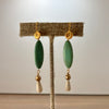 A pair of bohemian-style drop earrings displayed on a burlap-covered jewelry stand. The earrings feature a textured gold leaf-shaped top, a smooth olive green oval bead, and a small faceted gold bead suspending a cream-colored teardrop bead at the bottom.