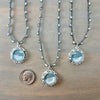 Three silver beaded necklaces with oval light blue cabochon pendants are displayed on a wooden surface, with a US quarter coin below the center pendant for scale.