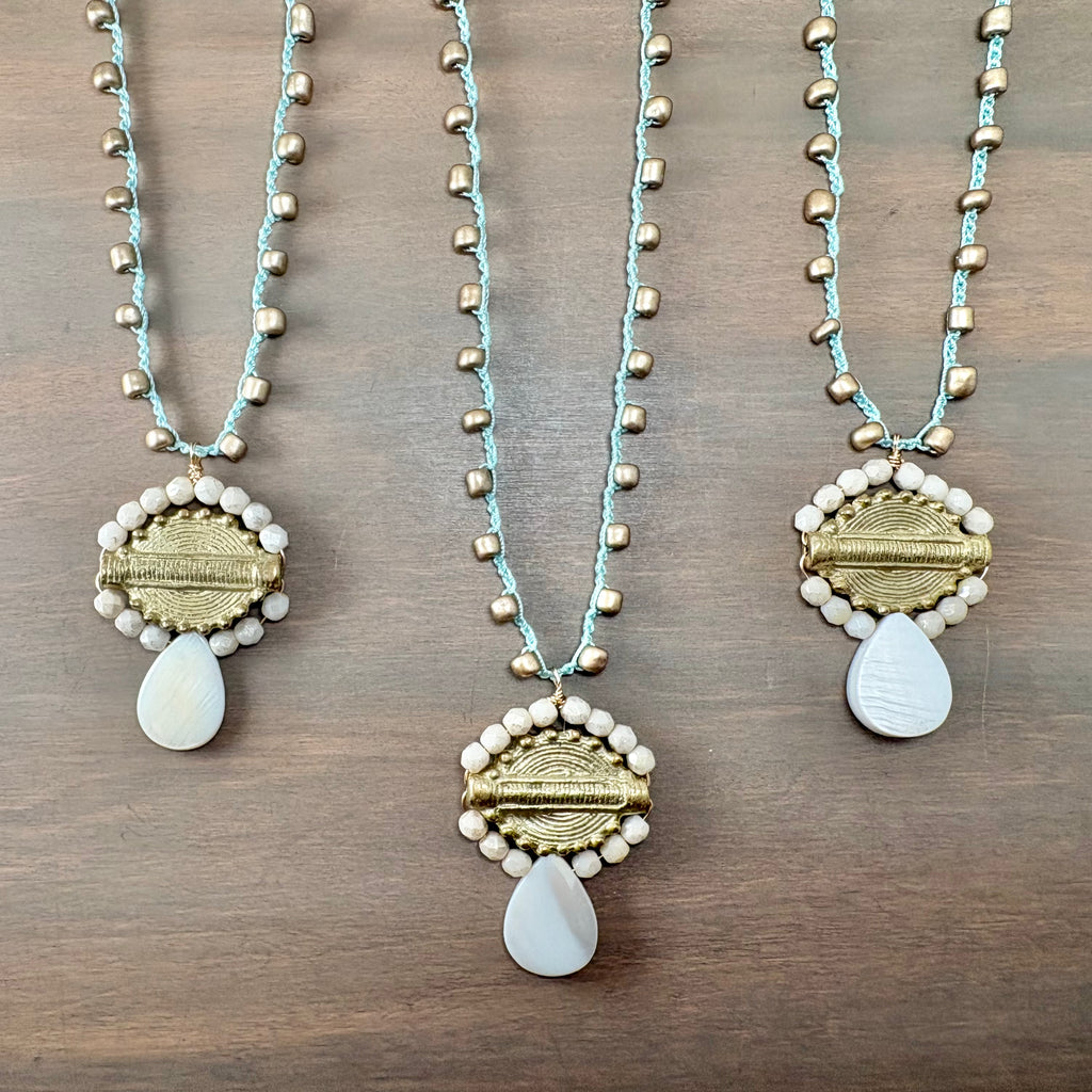 Three bohemian-style beaded necklaces displayed on a dark wood background. The necklaces feature light blue cords, metallic gold beads, white pearl-like beads, and a central gold textured oval with a white teardrop pendant.