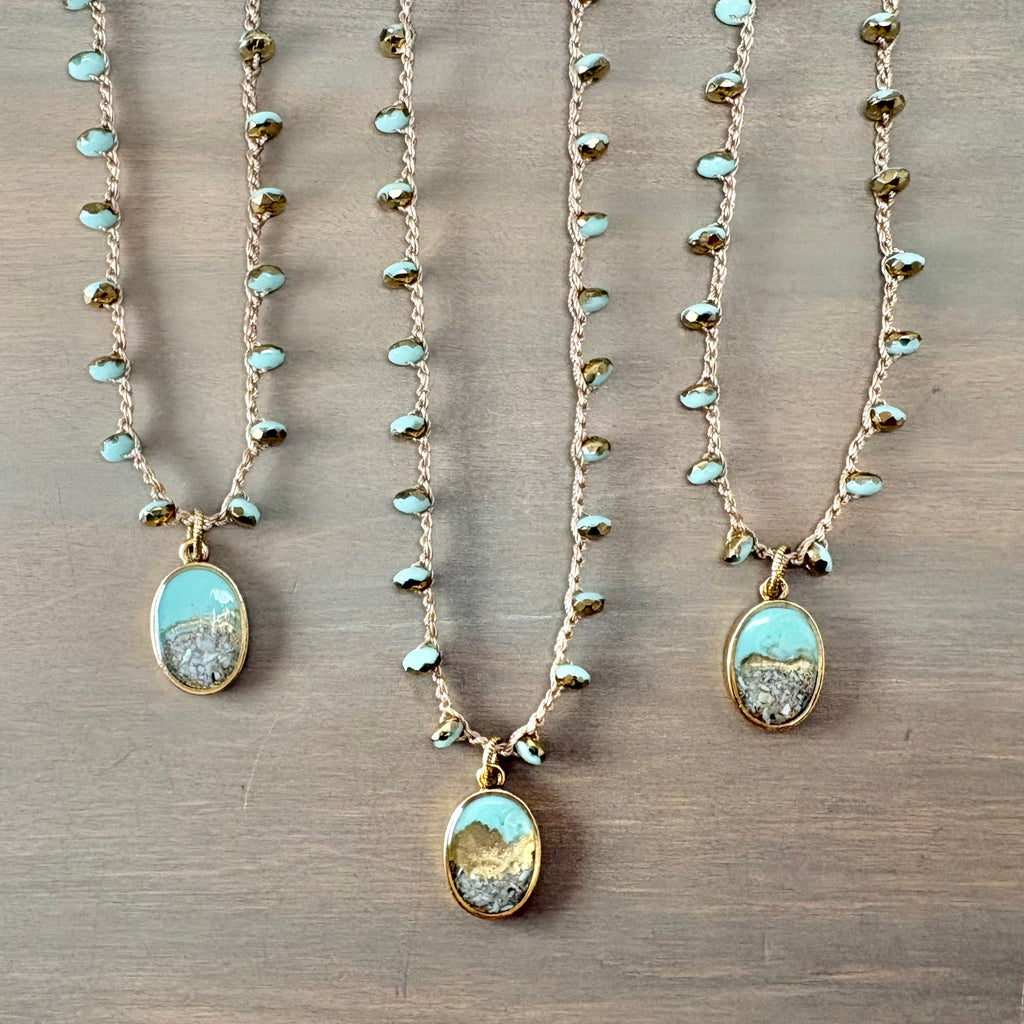 Three gold-toned beaded necklaces with oval pendants featuring a light blue and white landscape design, arranged on a brown wooden surface.