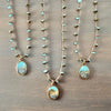 Three gold-toned beaded necklaces with oval pendants featuring a light blue and white landscape design, arranged on a brown wooden surface.