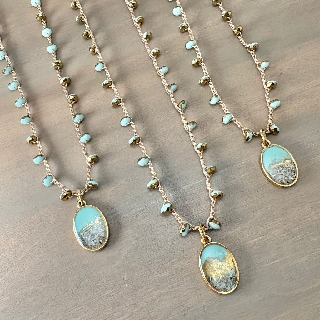 Three gold-toned beaded necklaces with oval turquoise and mountain landscape pendants, arranged diagonally on a gray wood background.