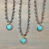 Three beaded necklaces with round turquoise pigment pendants and gold-toned frames, displayed on a gray wood background.