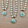 Three pearl bead necklaces with round gold-tone pendants featuring a blue and white wave design, displayed on a wooden surface.