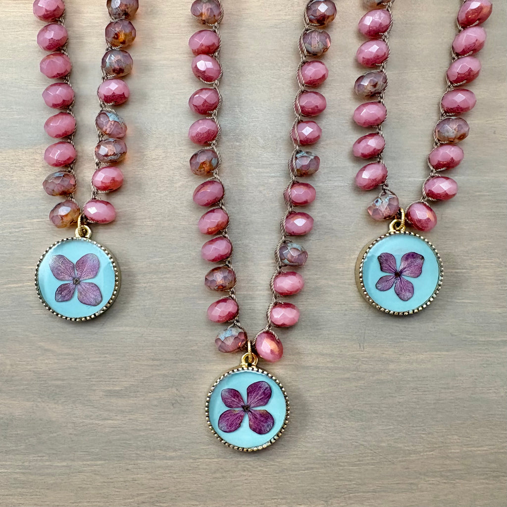 Three beaded necklaces with faceted pink beads and round light blue pampas grass pendant charms, arranged on a light wood background.