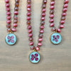 Three beaded necklaces with faceted pink beads and round light blue pampas grass pendant charms, arranged on a light wood background.