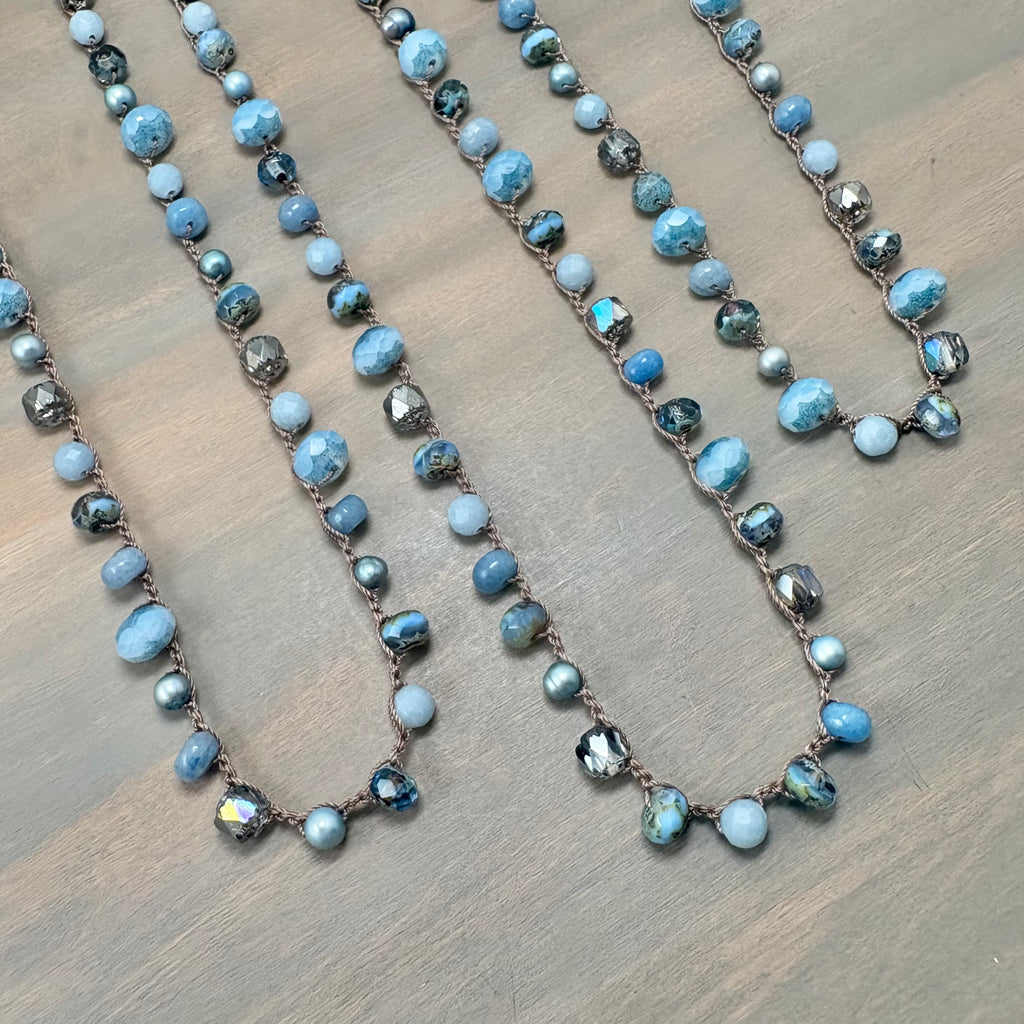 Three beaded necklaces with light blue, white, and dark gray marbled beads on a brown cord, arranged on a gray wood-grain surface.