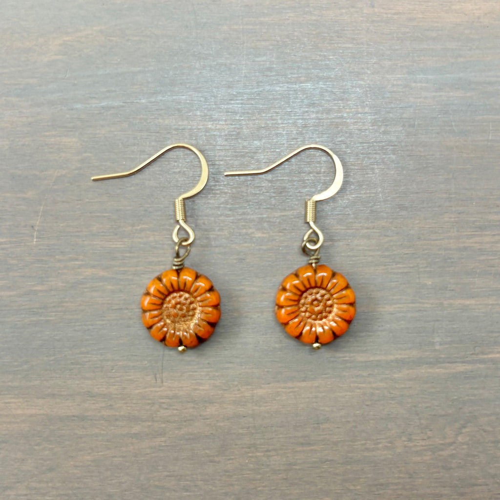 A pair of handmade dangle earrings featuring textured orange flower charms with gold-tone fishhook wires, displayed on a gray wood-grain surface.
