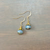 A pair of handmade dangle earrings featuring faceted light blue beads with gold-toned metal caps and fishhook wires, displayed on a dark wood surface.