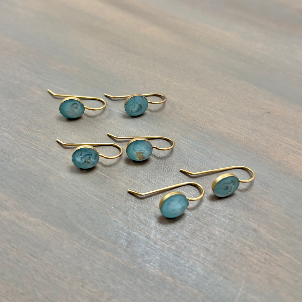 Six gold-toned ear hooks, each set with a small oval turquoise-blue stone, arranged on a gray wood-textured surface.