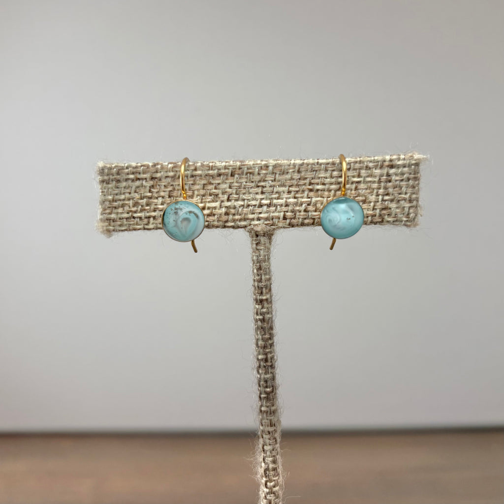 A pair of gold-tone drop earrings with round light blue marbled beads, displayed on a burlap-covered jewelry stand against a neutral gray background.