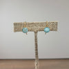 A pair of gold-tone drop earrings with round light blue marbled beads, displayed on a burlap-covered jewelry stand against a neutral gray background.