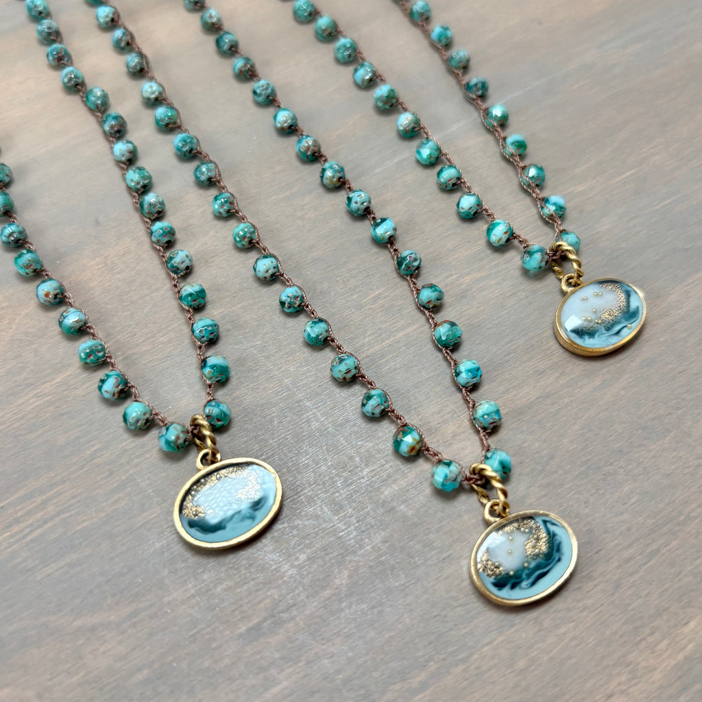 Three beaded necklaces with turquoise-blue marbled stone pendants and bronze-toned chains, arranged diagonally on a brown wooden surface.