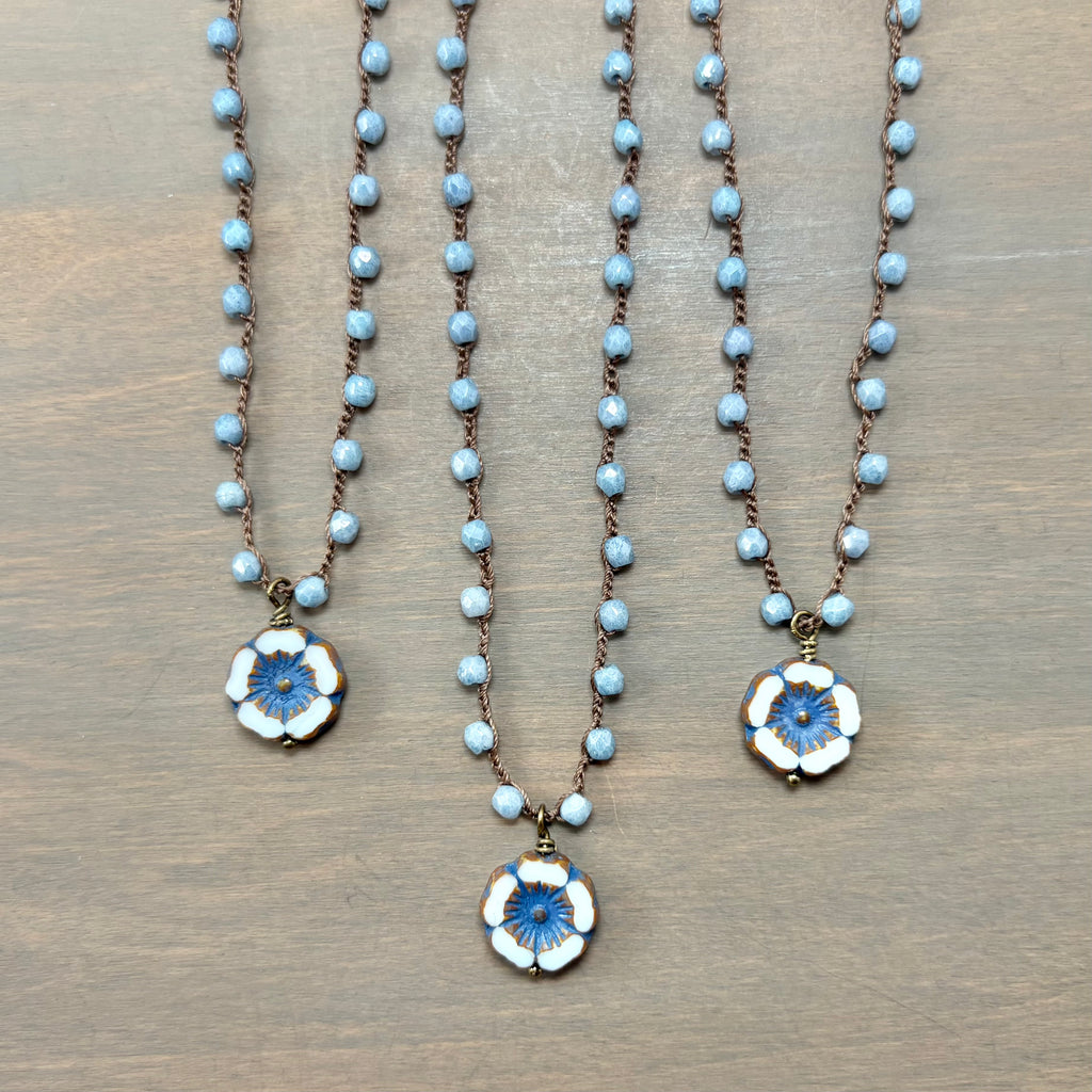 Three beaded necklaces with light blue beads and floral pendants in white, blue, and gold, arranged on a brown wooden surface.