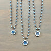 Three beaded necklaces with light blue beads and floral pendants in white, blue, and gold, arranged on a brown wooden surface.