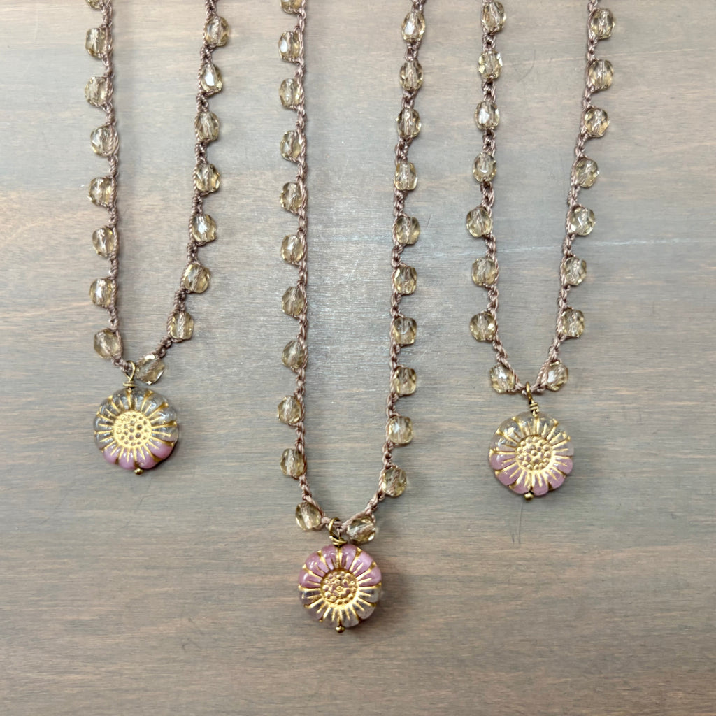 Three beaded necklaces with round daisy-like flower pendants in pink, gold, and white, displayed on a gray wood background.
