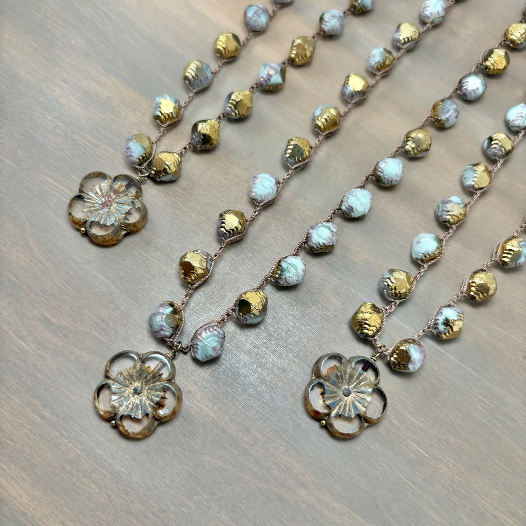Three strands of beaded jewelry laid out on a wooden surface, featuring textured gold-toned beads, iridescent light blue beads, and floral metal charms.