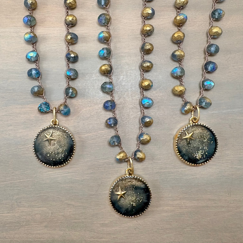 Three beaded necklaces with round galaxy-themed pendants are arranged on a light gray wooden surface. The necklaces feature faceted blue and gold beads, with pendants displaying a dark blue and black starry sky design with gold star details.