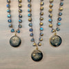 Three beaded necklaces with round galaxy-themed pendants are arranged on a light gray wooden surface. The necklaces feature faceted blue and gold beads, with pendants displaying a dark blue and black starry sky design with gold star details.