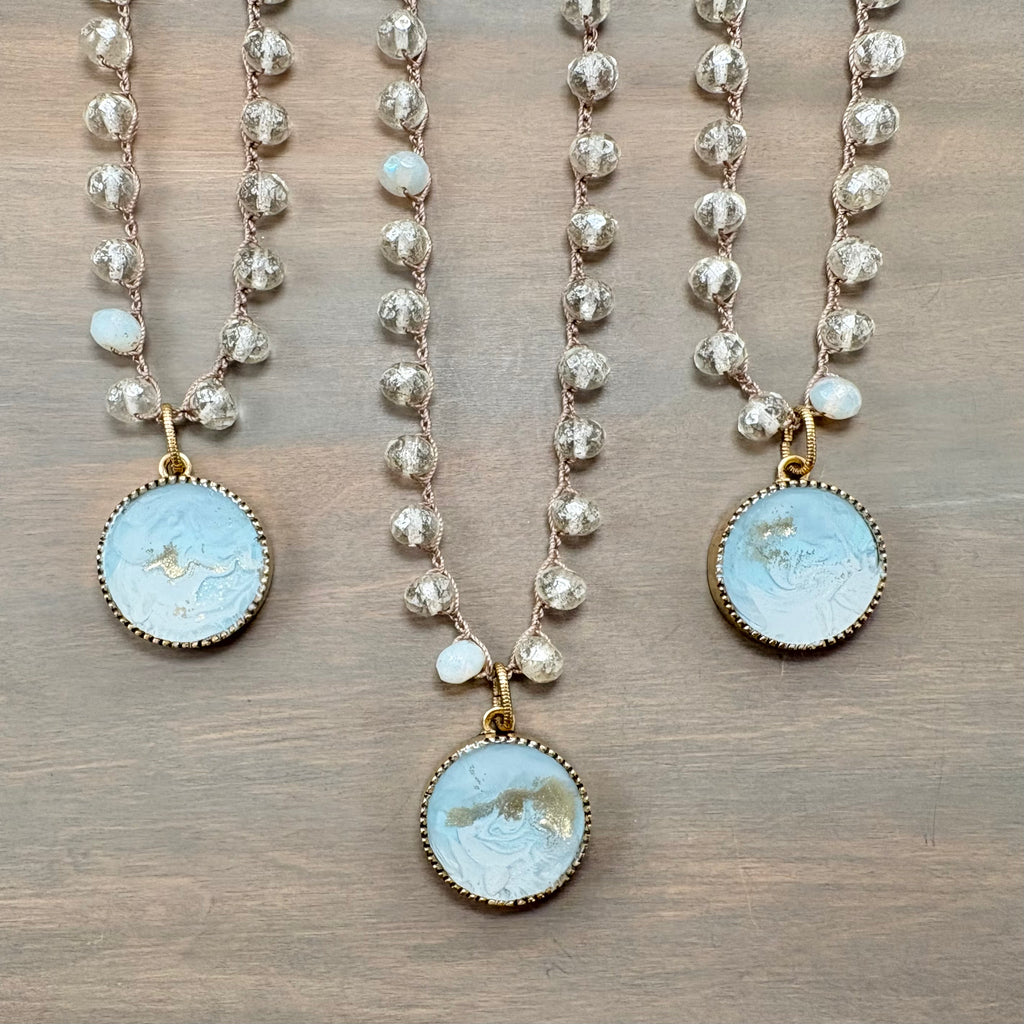 Three beaded necklaces with round light blue carved stone pendants and gold-toned settings, displayed on a wooden surface.