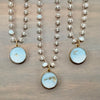 Three beaded necklaces with round light blue carved stone pendants and gold-toned settings, displayed on a wooden surface.