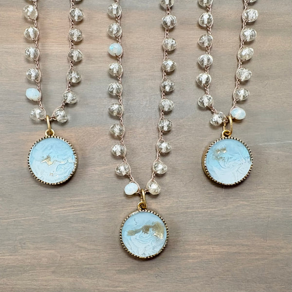 Three beaded necklaces with round light blue carved stone pendants and gold-toned settings, displayed on a wooden surface.