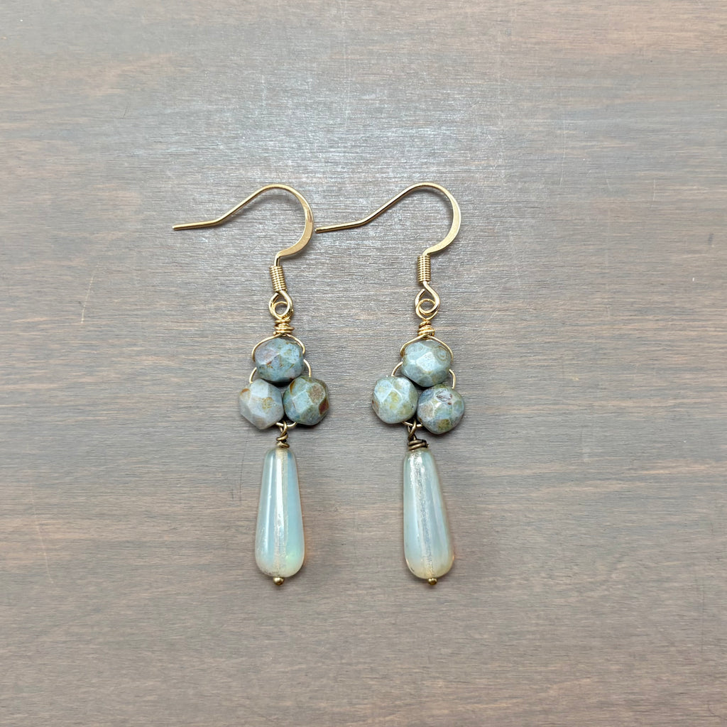 A pair of handmade dangle earrings featuring light blue faceted beads and teardrop-shaped iridescent white beads, suspended from gold-tone fishhook wires on a gray wood background.