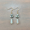 A pair of handmade dangle earrings featuring light blue faceted beads and teardrop-shaped iridescent white beads, suspended from gold-tone fishhook wires on a gray wood background.