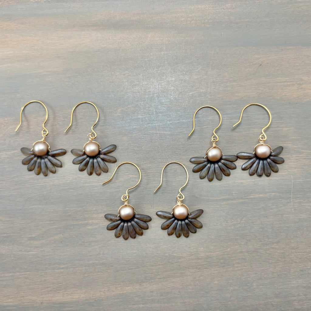 Five pairs of handmade daisy earrings with dark brown wooden petals, bronze metal hooks, and round pearl-like centers, arranged on a gray wooden surface.