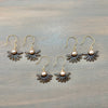 Five pairs of handmade daisy earrings with dark brown wooden petals, bronze metal hooks, and round pearl-like centers, arranged on a gray wooden surface.