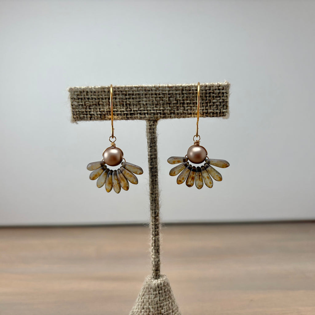 A pair of handmade daisy earrings with bronze-toned petals, a central brown pearl, and gold hooks, displayed on a burlap-covered jewelry stand against a neutral gray background.