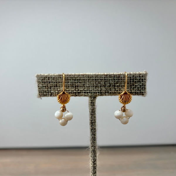 A pair of gold and white pearl dangle earrings displayed on a textured fabric jewelry stand against a plain light gray background.