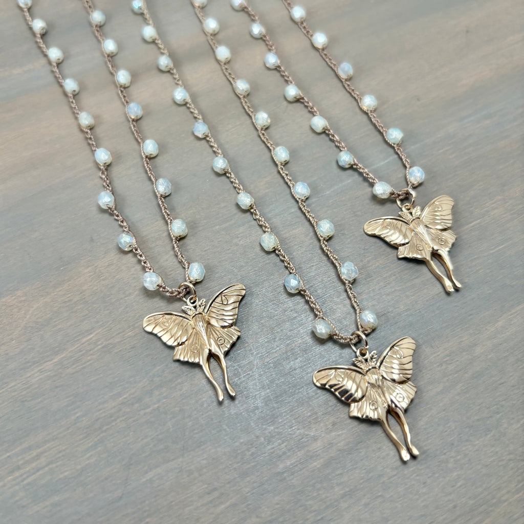 Three gold fairy luna moth necklaces with  small white pearl-like beads on a textured gray wooden surface.